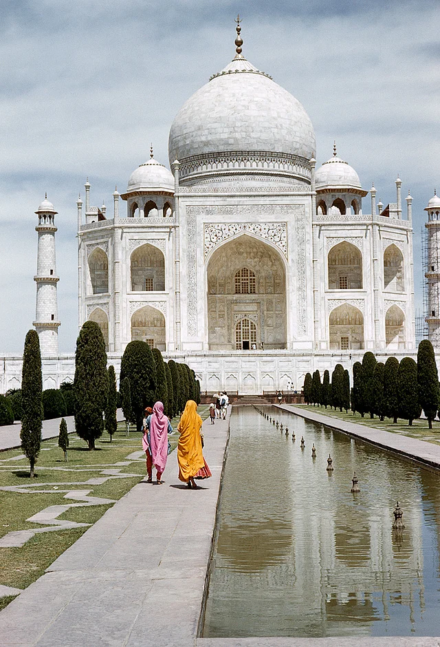 Women in bright dresses walk by a fountain with the Taj Mahal in the background, 1959.