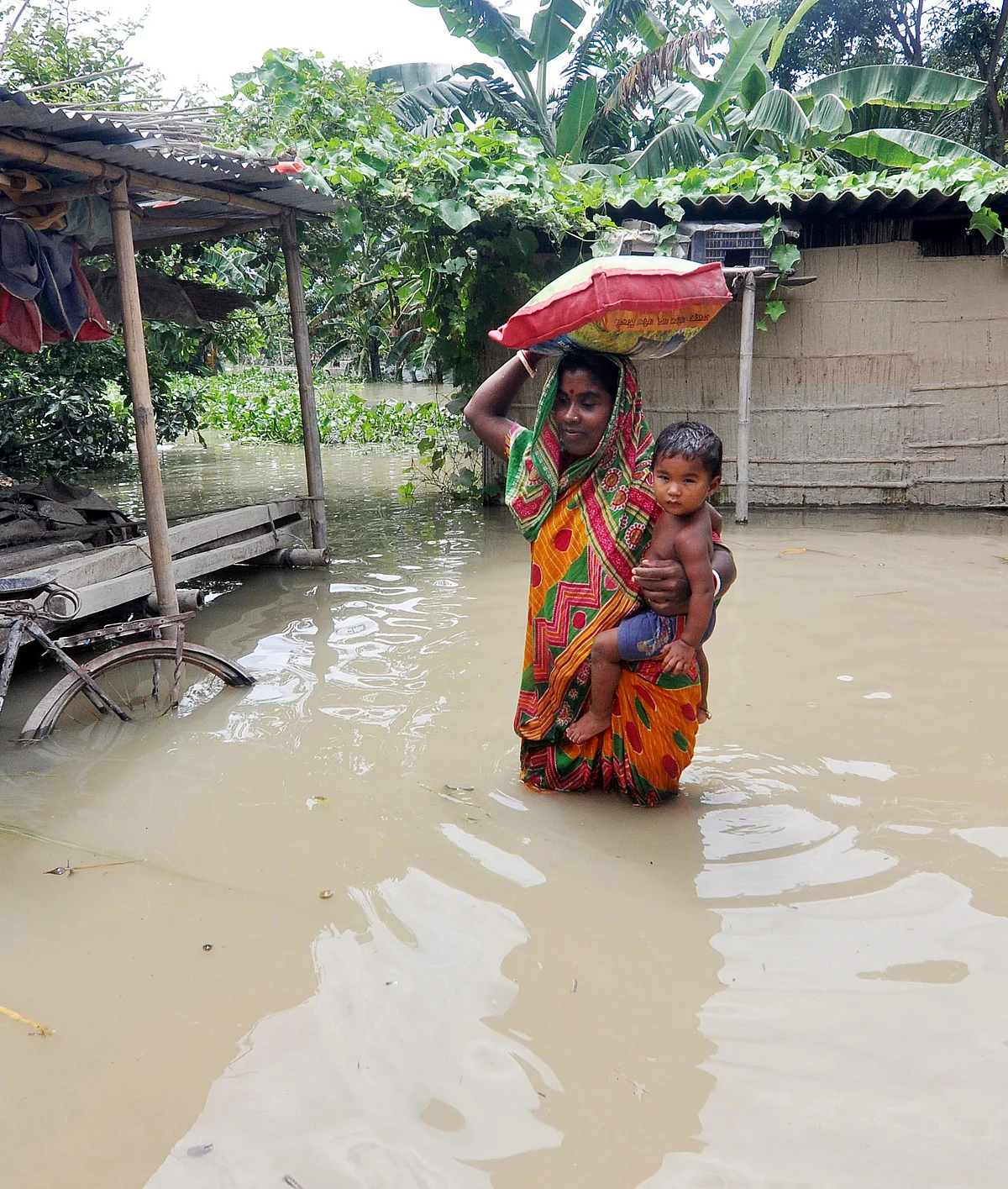 Assam, July 23 (ANI): A woman with her child stand in the flooded water of Murkata village in Morigaon district on Thursday.