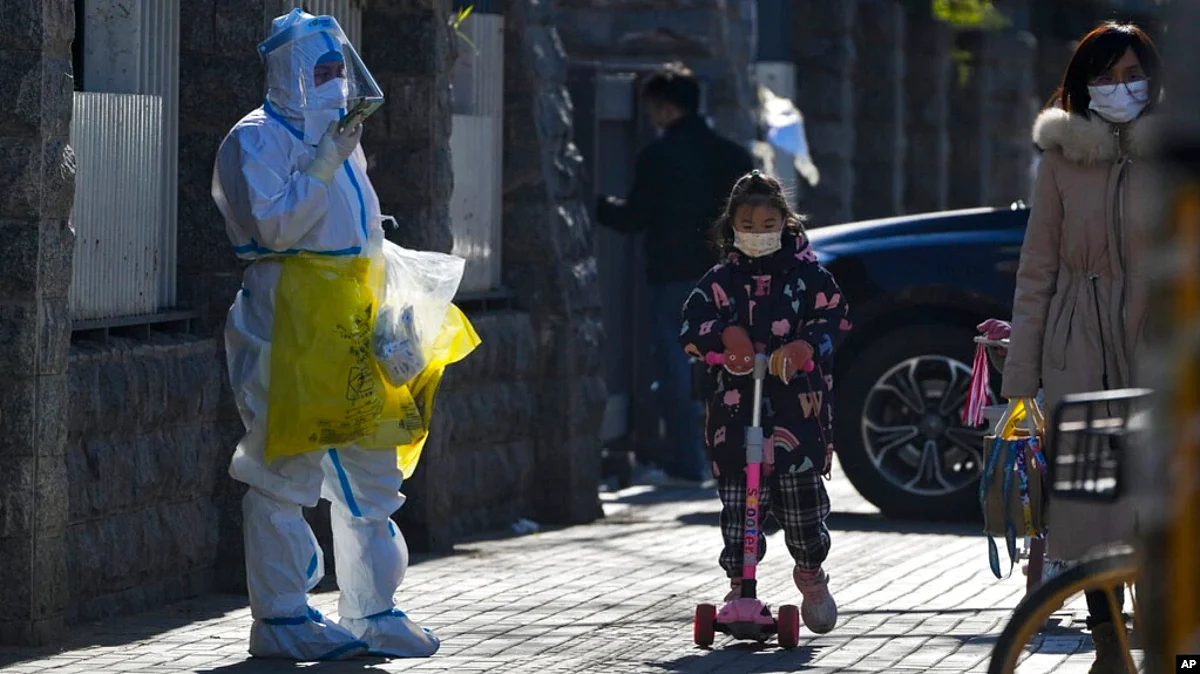 A child wearing a face mask and riding on a scooter passes by a worker in protective suit on his way to collect COVID samples from the lockdown residents in Beijing, Dec. 1, 2022. (AP)