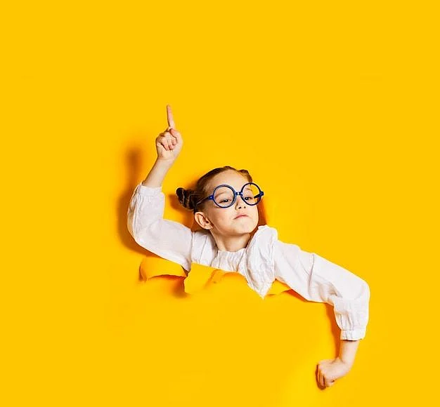 Premium Photo Pretty smart looking schoolgirl in glasses breaking through torn in yellow paper wall holding her index finger up little teacher to be teaching how to live and behaive new rules of live concept