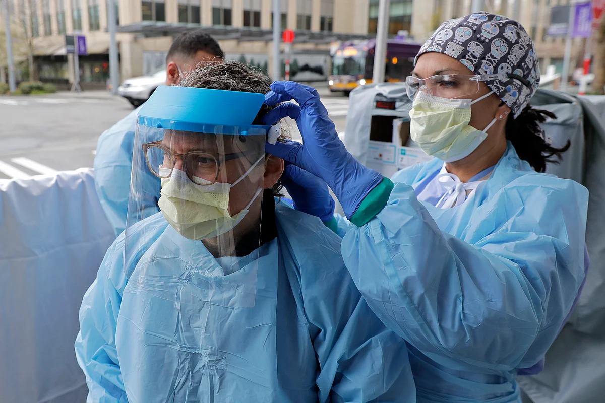 An emergency services nurse helps a colleague put on a medical face shield.